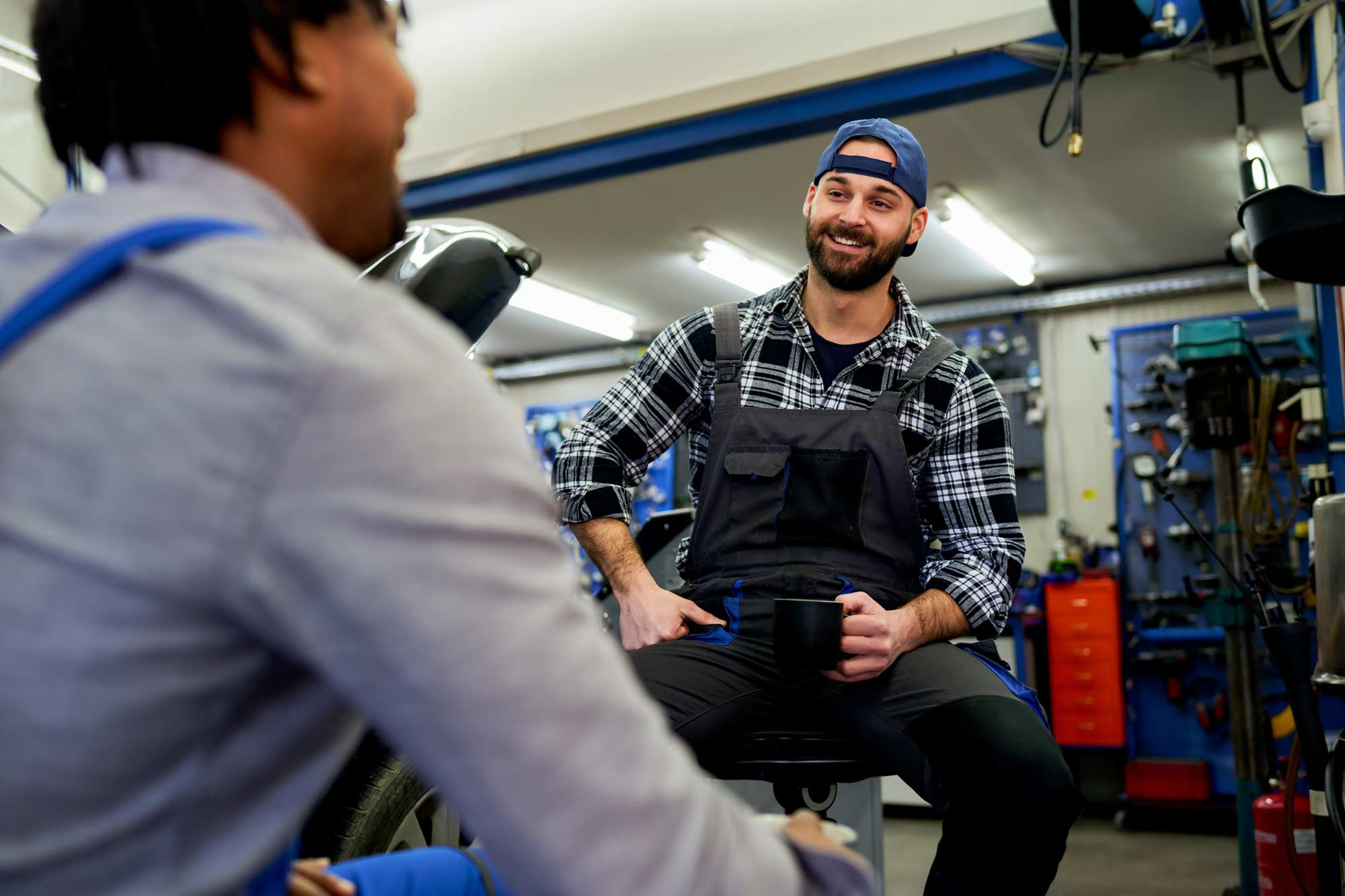 Two-cheerful-auto-mechanics-are-taking-a-coffee-break-and-chatting-together-in-their-garage,-enjoying-a-moment-of-camaraderie-amidst-their-workday-web